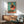 May include: Framed poster showing a stylized rooster with yellow head and red comb wearing a black polka dot dress, standing on a vinyl record turntable against a teal background with coral circle. Displayed above a wooden sideboard with white ceramic lamp in a mid-century modern living room with green upholstered chair.
