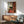 May include: Framed reproduction of Modigliani's elongated portrait showing a woman in dark green top and coral red skirt seated in wooden chair, displayed above mid-century modern wood credenza with ceramic table lamp, next to sage green upholstered armchair in sunlit room with white walls.