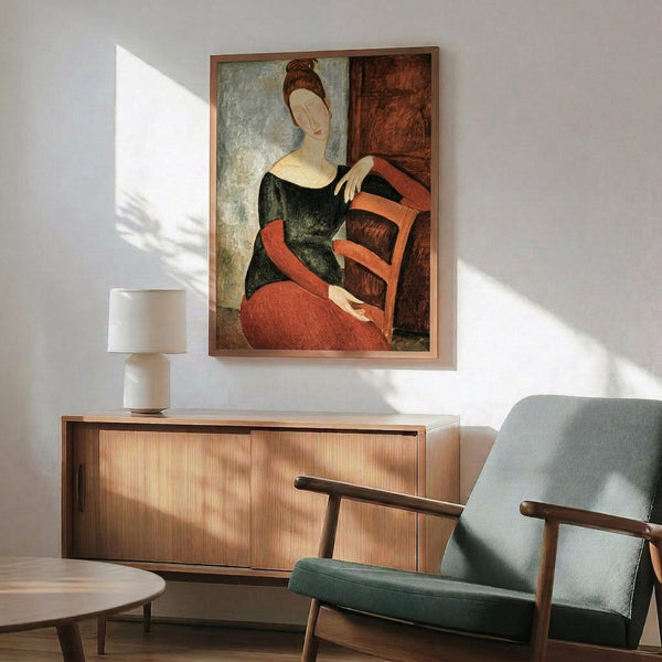 May include: Framed reproduction of Modigliani's elongated portrait showing a woman in dark green top and coral red skirt seated in wooden chair, displayed above mid-century modern wood credenza with ceramic table lamp, next to sage green upholstered armchair in sunlit room with white walls.