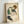 May include: Framed reproduction of an expressionist drawing showing a seated woman with curly reddish hair wearing a green sleeveless top and dark stockings. The artwork features loose, sketchy lines and muted colors on cream paper, displayed in a light wood frame leaning against a white wall.