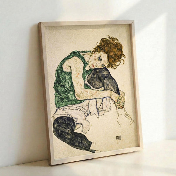 May include: Framed reproduction of an expressionist drawing showing a seated woman with curly reddish hair wearing a green sleeveless top and dark stockings. The artwork features loose, sketchy lines and muted colors on cream paper, displayed in a light wood frame leaning against a white wall.