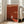 May include: A framed minimalist art poster with rust-red background displayed in a bright living room corner. The poster shows a silhouetted figure standing before a white doorway or opening, with geometric shapes and typography. The wooden frame leans against white walls near tall windows, positioned next to a beige upholstered chair. Natural sunlight streams across the wooden floor, creating shadows and highlighting the poster's warm earth tones and clean modern design.