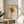May include: Framed poster featuring abstract floral line drawing with orange and yellow circular blooms on cream background, displayed on round wooden stool beside beige upholstered chair. Natural sunlight creates geometric shadows on white wall. Green plant leaves visible in corner. Print shows minimalist botanical illustration in warm autumn tones with black linear stems and leaves.