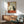 May include: Framed poster artwork in wood frame hanging on white wall above mid-century modern wooden credenza with white table lamp. Green upholstered armchair with wooden arms visible in foreground. Artwork shows black cat holding wine glass at table with wine bottle, grapes and cheese in traditional Japanese ukiyo-e style with beige background and red Japanese text cartouche. Warm natural lighting creates shadows on wall.