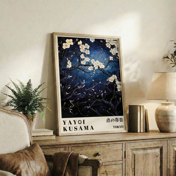May include: Framed poster with deep blue night scene featuring white cherry blossoms on dark branches against starry sky, displayed on rustic wooden console table beside cream armchair, fern plant, ceramic vases and stacked books in neutral-toned living room setting