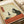 Japanese woodblock print style artwork featuring a black cat sitting on a red table, holding a wine glass. The cat has detailed whiskers and pointed ears. On the table are dark grapes, a wine bottle with a label, and what appears to be cheese. The background is cream colored with Japanese characters in a red rectangular cartouche in the upper left corner. The print has an aged, vintage appearance with subtle texture and warm tones throughout.