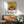 May include: Framed poster showing a colorful watermelon illustration with Japanese text, displayed above a wooden credenza with white lamp in a modern living room setting. The artwork features a whole watermelon on yellow dotted background and watermelon slice on blue and green surface. Mid-century modern green armchair with wooden arms sits nearby with natural sunlight streaming across white walls.