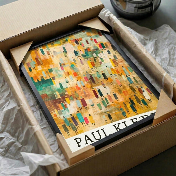 May include: Framed abstract art print in black frame being unpacked from cardboard shipping box with tissue paper. Artwork features colorful geometric pattern with small figure-like shapes in warm oranges, yellows, greens, and earth tones. White label with black text visible at bottom of frame. Surrounded by brown packaging materials on wooden surface.