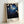 Framed poster leaning against white wall featuring nighttime scene with white cherry blossoms on dark blue starry background. Text at bottom reads "YAYOI KUSAMA" in black letters and Japanese characters with "TOKYO" in smaller text. Artwork shows flowing branches with delicate white flowers scattered across deep blue to black gradient sky filled with small white dots resembling stars. Natural wood frame surrounds the print.