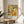 May include: A framed art print displaying an abstract composition with colorful geometric figures resembling people in warm yellows, oranges, teals, and earth tones. The poster features "PAUL KLEE" text at bottom with "PLAYFUL ABSTRACTION MODERNISM 1879-1940" in smaller print. The thin wooden frame rests on a light wood round side table next to a beige upholstered chair, with plant leaves and natural window light creating shadows on white walls.
