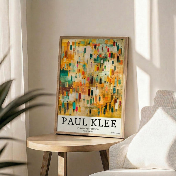 May include: A framed art print displaying an abstract composition with colorful geometric figures resembling people in warm yellows, oranges, teals, and earth tones. The poster features "PAUL KLEE" text at bottom with "PLAYFUL ABSTRACTION MODERNISM 1879-1940" in smaller print. The thin wooden frame rests on a light wood round side table next to a beige upholstered chair, with plant leaves and natural window light creating shadows on white walls.