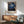 May include: Framed art print featuring cherry blossoms against deep blue night sky with white dots, displayed on white wall above wooden sideboard with lamp. Mid-century modern sage green armchair with wooden arms sits nearby. Natural sunlight creates soft shadows across the minimalist interior scene.