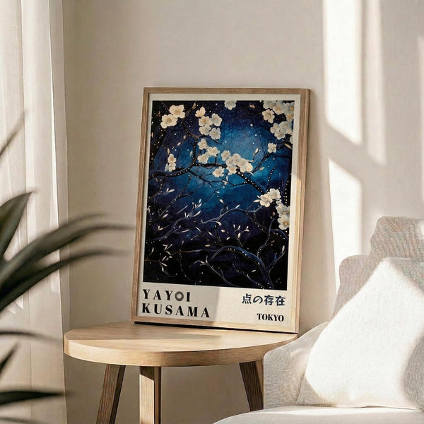 May include: Framed art print featuring a dreamy night sky scene with white cherry blossoms against deep blue starry background, displayed on a light wood round side table next to a beige upholstered chair. Natural sunlight streams across the white wall creating geometric shadows. The artwork shows delicate white flowers on dark branches with scattered stars and cosmic blue tones, presented in a thin natural wood frame.
