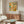 May include: Modern living room featuring colorful abstract artwork with geometric patterns in yellow, orange, green, and blue tones displayed above a mid-century walnut credenza. White upholstered chair and glass-top coffee table complete the minimalist Scandinavian-style interior with cream walls and natural lighting.