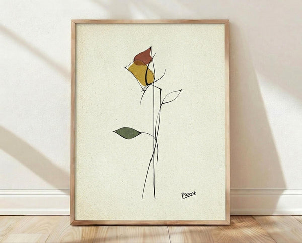 May include: A framed minimalist line drawing of a single rose with geometric colored petals in yellow, brown, and red tones, plus a green leaf on a long black stem. The artwork sits on light wood flooring against a white textured wall with natural lighting casting soft shadows. The piece is displayed in a simple light wood frame on cream-colored paper with a signature in the bottom right corner.