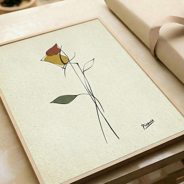 May include: Framed minimalist line art of a single rose with geometric colored petals in red, yellow, and green on cream textured paper. The delicate black line drawing shows a long stem with leaves, displayed in a light wood frame on a neutral surface with soft natural lighting and gift wrapping materials visible nearby.
