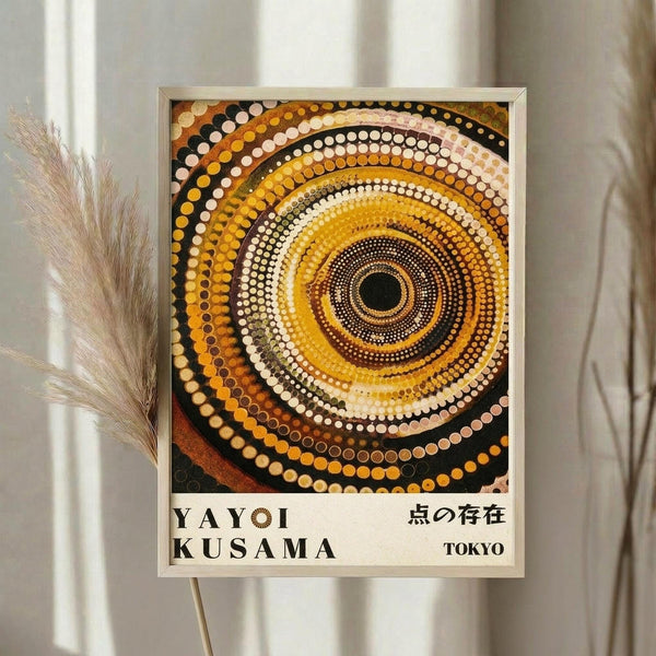 May include: Framed poster with concentric circles in warm earth tones - yellows, oranges, browns, and cream dots radiating from a dark center. The dotted pattern creates a mesmerizing spiral effect. Text at bottom reads "YAYOI KUSAMA" and Japanese characters with "TOKYO". Light wood frame hangs on white wall next to dried pampas grass stems, creating a minimalist gallery wall aesthetic.