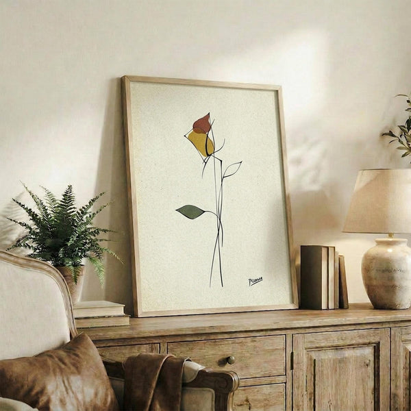May include: Framed minimalist line art print featuring a single rose with yellow and burgundy petals and simple black stem, displayed on rustic wooden sideboard alongside fern plant, ceramic vase, stacked books, and brown leather chair in warm-toned living room setting