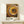 May include: Framed poster with concentric circles in warm earth tones - yellows, oranges, browns, and cream dots radiating outward from dark center, reminiscent of Aboriginal dot painting style. Text reads "YAYOI KUSAMA" and Japanese characters with "TOKYO" below. Light wooden frame sits on pale wooden shelf against white wall, with small white wrapped gift box tied with twine and dried sprig beside it.