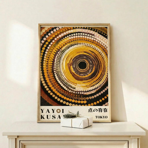 May include: Framed poster with concentric circles in warm earth tones - yellows, oranges, browns, and cream dots radiating outward from dark center, reminiscent of Aboriginal dot painting style. Text reads "YAYOI KUSAMA" and Japanese characters with "TOKYO" below. Light wooden frame sits on pale wooden shelf against white wall, with small white wrapped gift box tied with twine and dried sprig beside it.