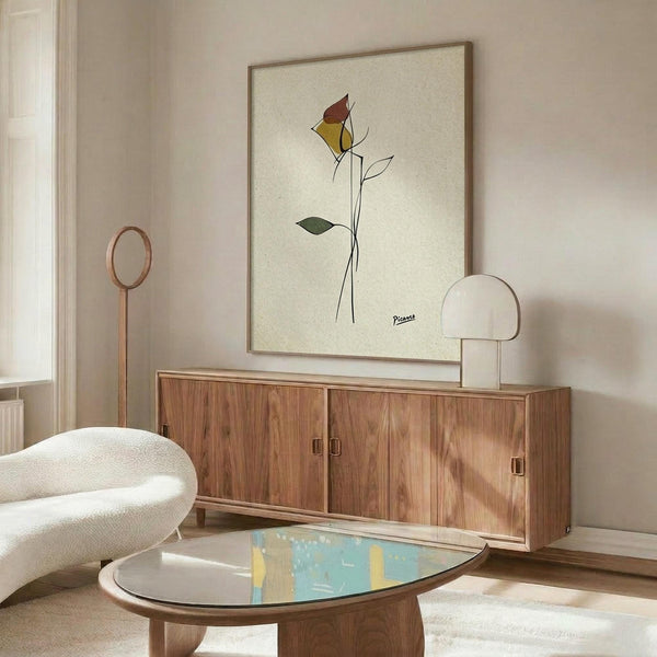 May include: Framed minimalist line drawing of a rose with colorful geometric petals in red, yellow, and green on cream background, displayed above wooden sideboard in modern living room with white curved chair, glass coffee table, and warm natural lighting