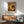 May include: Interior scene showing a framed poster with concentric circles in warm tones of yellow, orange, brown and white dots creating a mesmerizing spiral pattern. The artwork hangs above a wooden sideboard with clean lines, next to a modern green upholstered chair with wooden arms. A white ceramic table lamp and round wooden coffee table complete the mid-century modern room setting with white walls and natural lighting.