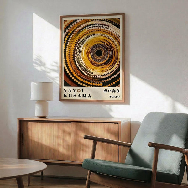 May include: Interior scene showing a framed poster with concentric circles in warm tones of yellow, orange, brown and white dots creating a mesmerizing spiral pattern. The artwork hangs above a wooden sideboard with clean lines, next to a modern green upholstered chair with wooden arms. A white ceramic table lamp and round wooden coffee table complete the mid-century modern room setting with white walls and natural lighting.