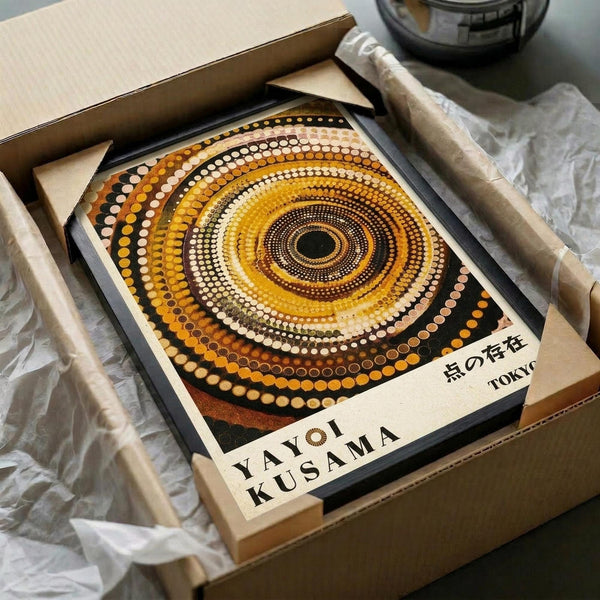 May include: Framed poster with concentric circles in warm earth tones - yellows, oranges, browns, and creams - created from dotted patterns radiating from center. Japanese text visible at bottom. Print sits in cardboard shipping box with protective tissue paper, surrounded by brown paper wrapping materials on white surface.