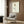 May include: Framed minimalist line art print showing a single stylized rose with brown, yellow and green geometric petals on cream background, displayed above a beige upholstered armchair with geometric pillow in modern living room with wood paneling, gold floor lamp, and dark side table with open book.