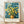 May include: Framed poster print displaying Van Gogh's painting of yellow wildflowers in a brown ceramic vase against a swirling blue background, featuring museum exhibition text below the artwork. The light wood frame leans against a white wall on wooden flooring with natural shadows cast across the scene.