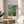 May include: Framed Van Gogh museum poster featuring yellow flowers in a vase against blue brushstroke background, displayed on round wooden side table next to beige upholstered chair with white throw pillow, natural lighting from window creating soft shadows on neutral wall with plant leaves visible in corner.