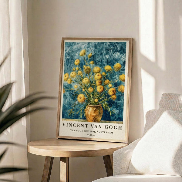 May include: Framed Van Gogh museum poster featuring yellow flowers in a vase against blue brushstroke background, displayed on round wooden side table next to beige upholstered chair with white throw pillow, natural lighting from window creating soft shadows on neutral wall with plant leaves visible in corner.