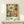 May include: Framed botanical art print featuring large cream and orange flowers with detailed petals and green foliage against a dark olive background with dotted patterns. The artwork displays "YAYOI KUSAMA" and "Modern Art 1940 TOKYO" text at bottom. Print sits in light wood frame on white wooden shelf next to small white gift box with greenery sprig, against cream wall with soft natural lighting.