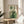May include: Framed botanical abstract art print with green geometric shapes, plants, and striped rainbow arch displayed on light wood round side table next to beige upholstered chair. Sunlight casts shadows across white walls in minimalist living room setting with potted plant visible in corner.