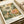 A vintage-style botanical art print featuring large detailed flowers in cream, orange, and pink tones against a dark green background with dotted patterns. The artwork shows Japanese-inspired floral illustrations with intricate line work. Text at bottom reads "YAYOI KUSAMA" and "Melody of the Sea Breeze" with "Modern Art 1940" and "TOKYO" labels. A color palette strip appears on the right side. The print lies flat on white paper with cream ribbon nearby.