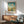 May include: Vintage-style Georgia travel poster featuring Savannah Historic District with classic buildings and cloudy blue sky, displayed in wooden frame above mid-century modern credenza with white ceramic lamp, alongside sage green upholstered armchair with wooden arms in sunlit room with white walls