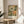 May include: Framed botanical art print with vintage-inspired floral design featuring large blooms in green, orange, and cream colors with dotted patterns. The artwork sits on a round wooden side table next to a beige upholstered chair, with natural sunlight casting shadows and a plant visible in the corner of the neutral-toned room.