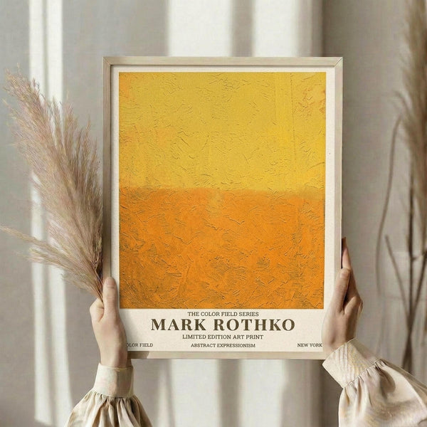 May include: Person holding a framed art print featuring a yellow to orange gradient color field painting with textured brushstrokes, displayed against white curtains with dried pampas grass visible to the left side.