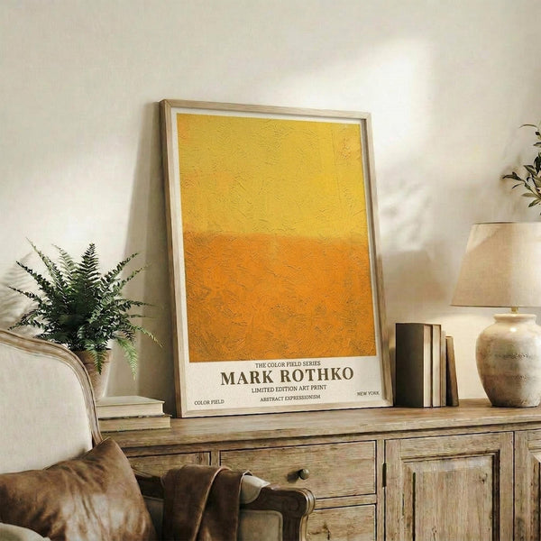 May include: Framed abstract art print with yellow and orange color field design in warm gradient tones, displayed on rustic wooden sideboard alongside fern plant, ceramic vases, and books in neutral-toned living room with cream chair and textured throw blanket.