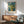 May include: Van Gogh yellow flower artwork in wooden frame hanging on white wall above wooden sideboard with white lamp. Green upholstered armchair with wooden arms sits nearby alongside round wooden coffee table. Sunlight creates geometric shadows across the minimalist room setting.