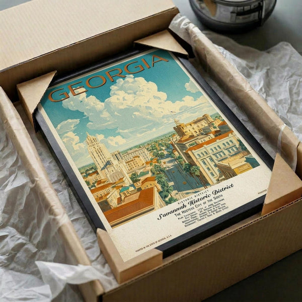 May include: Vintage-style travel poster of Georgia featuring Savannah Historic District with colonial buildings, Spanish moss-draped trees, and cloudy blue sky, displayed in black frame inside cardboard shipping box with protective tissue paper wrapping