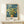 May include: Framed Van Gogh poster print featuring yellow chrysanthemum flowers in a brown vase against swirling blue brushstroke background, displayed on light wood console table with small white gift box, neutral wall backdrop, natural lighting creating soft shadows
