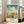 May include: Vintage-style travel poster featuring Savannah Historic District with turquoise sky, white puffy clouds, and colorful historic buildings with red tile roofs and palm trees. The poster shows "GEORGIA" in large orange letters at top and descriptive text at bottom. Framed in light wood and displayed in bright living room with beige sofa, white walls, and tall windows with natural light casting shadows on hardwood floors.