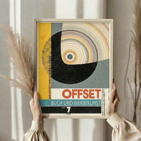 May include: Person holding framed vintage Bauhaus poster in light wood frame against neutral curtained background, with dried pampas grass visible to left. Artwork features bold geometric design with yellow, gray, black and teal color blocks, concentric circles, and German text reading "OFFSET BUCH UND WERBEKUNST" dated 1926. Mid-century modern aesthetic with textured, weathered appearance.