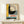 May include: Framed vintage Bauhaus poster featuring bold geometric design with yellow, black, and gray sections, concentric circles resembling a vinyl record, and German text "OFFSET BUCH UND WERBEKUNST". Wood frame sits on light wooden surface beside small wrapped gift with natural twine and dried sprig against cream wall.