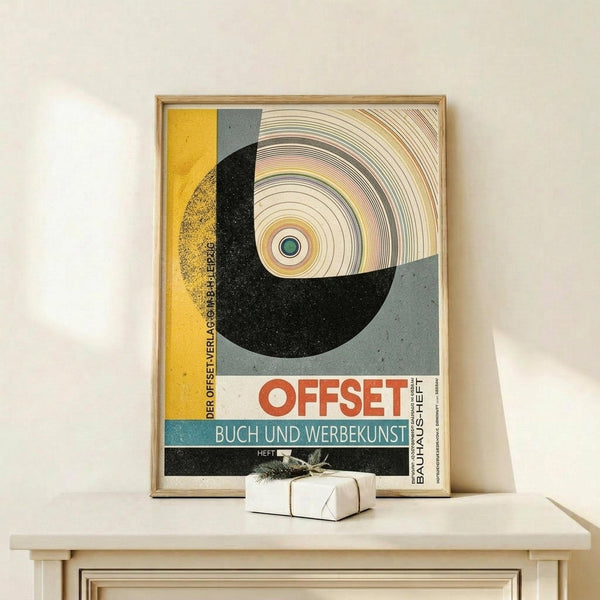 May include: Framed vintage Bauhaus poster featuring bold geometric design with yellow, black, and gray sections, concentric circles resembling a vinyl record, and German text "OFFSET BUCH UND WERBEKUNST". Wood frame sits on light wooden surface beside small wrapped gift with natural twine and dried sprig against cream wall.