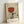 May include: Framed art print leaning against white wall showing abstract illustration with hands holding wine glass filled with red liquid, stylized eyes, and French text "Plaisir des yeux" at bottom. Artwork features muted beige, terracotta, and olive green colors in minimalist style within light wood frame.