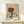 May include: Framed vintage-style poster with abstract illustration showing wine glass filled with red liquid, stylized hands, and eyes in muted earth tones on beige background, displayed on light wood shelf beside small white wrapped gift with greenery sprig against cream wall