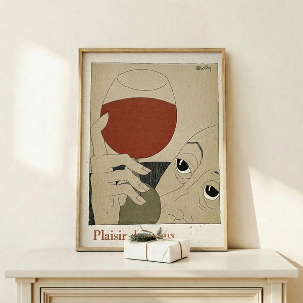 May include: Framed vintage-style poster with abstract illustration showing wine glass filled with red liquid, stylized hands, and eyes in muted earth tones on beige background, displayed on light wood shelf beside small white wrapped gift with greenery sprig against cream wall