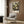 May include: Botanical art poster featuring circular flower and plant specimens in cream, yellow, and orange tones against a dark background, displayed in light wood frame above beige upholstered armchair with geometric patterned pillow, brass floor lamp, and dark round side table with open book in modern living room with wood paneled walls